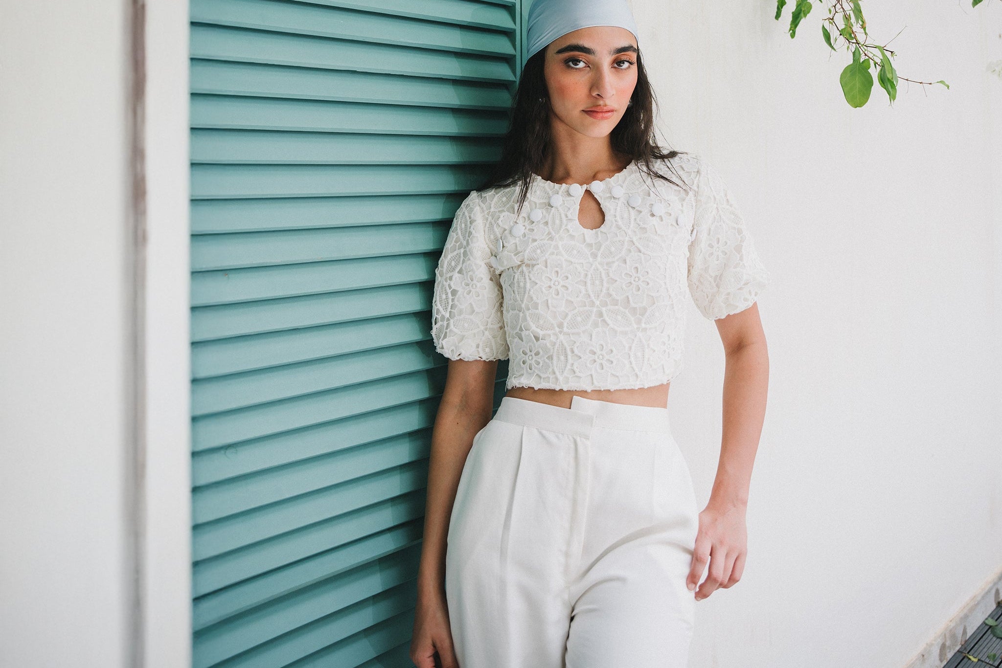 Woman wearing a light blue headscarf and white outfit standing in front of a teal shutter.